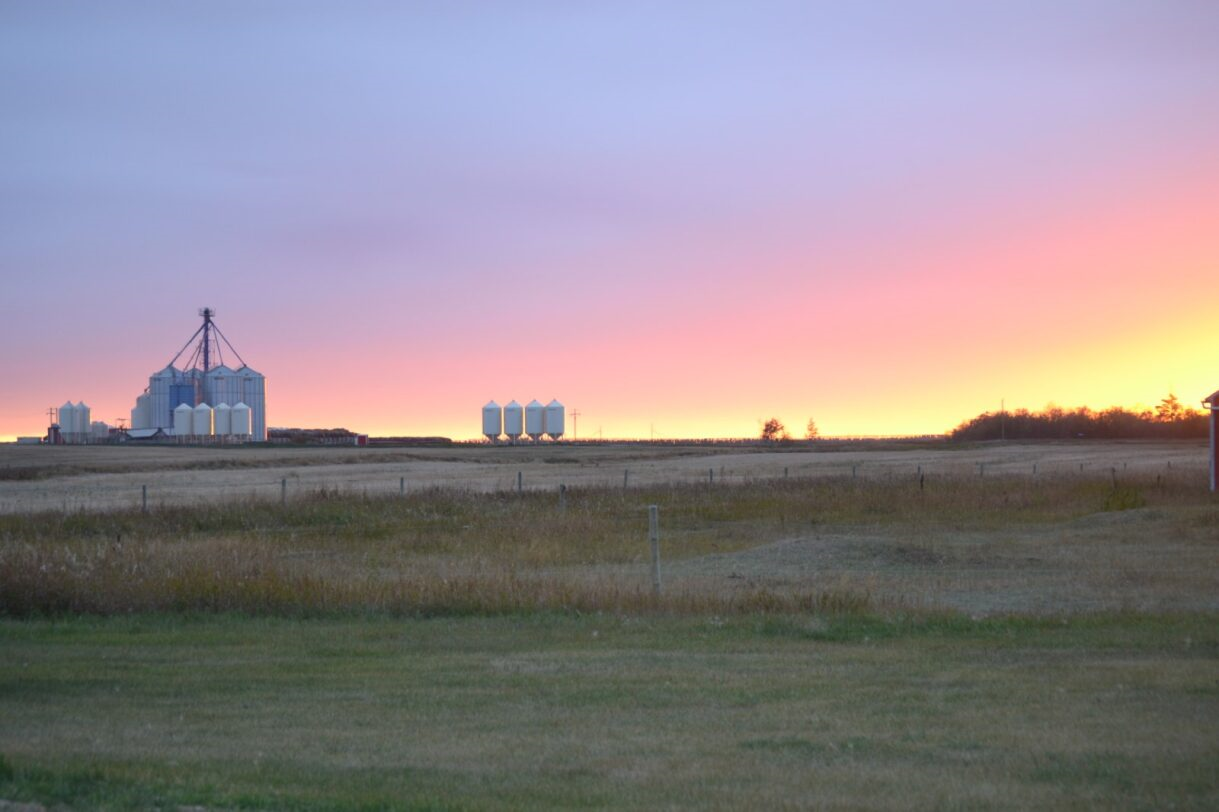 Sunset over a prairie landscape with grain silos in the distance, vibrant orange and pink sky.