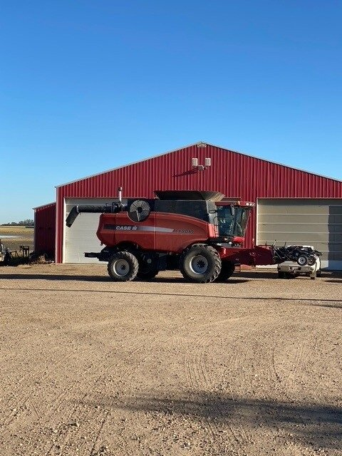 Red combine harvester in front of a red barn with a blue sky.