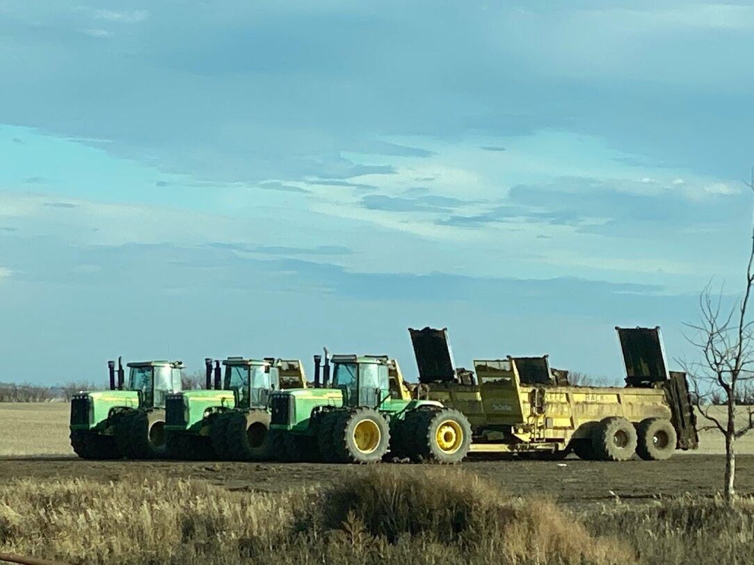 Three green tractors and a yellow manure spreader sit in a field under a cloudy sky.
