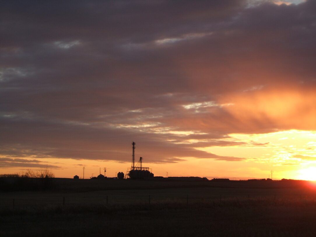 Silhouette of an oil rig against a sunset sky with orange and dark gray clouds.