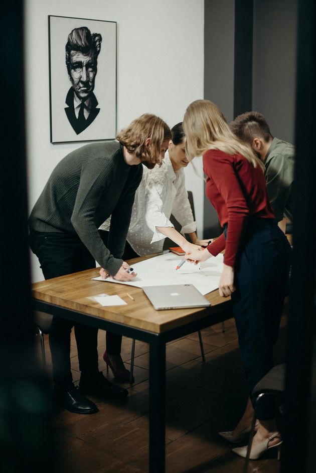 Vier collega's werken samen rond een houten tafel in een kantoor en bekijken documenten, met een portret aan de muur.