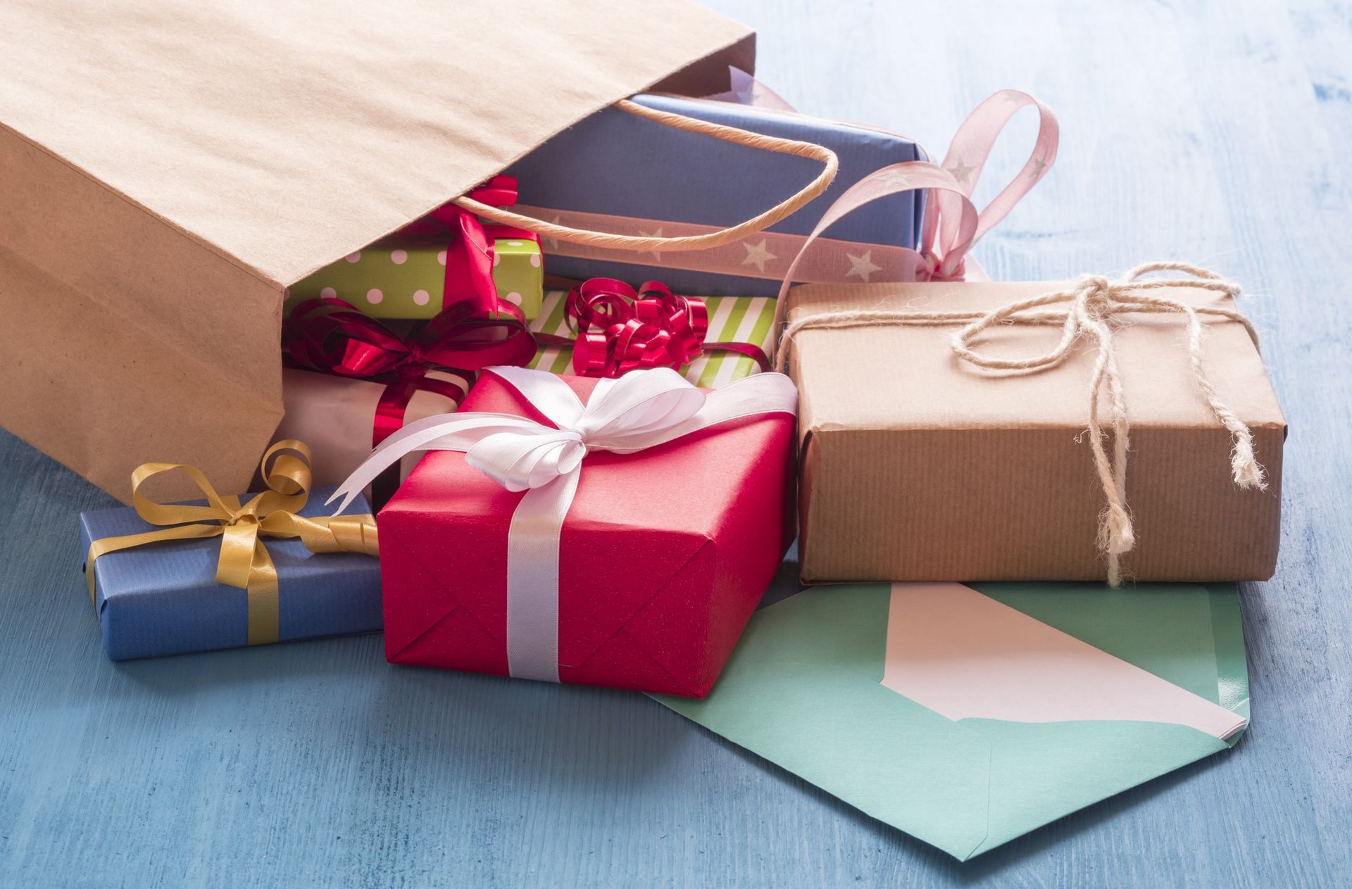 Gifts overflowing from a brown paper bag, including wrapped boxes in red, blue, and brown, with ribbons and an envelope on a blue surface.