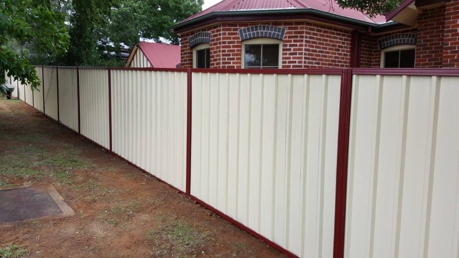 A Tall Cream-colored Metal Fence With Red Posts Lines The Perimeter Of A Brick House With A Red Roof — Anne & Ron Johnson Fencing in Werris Creek, NSW