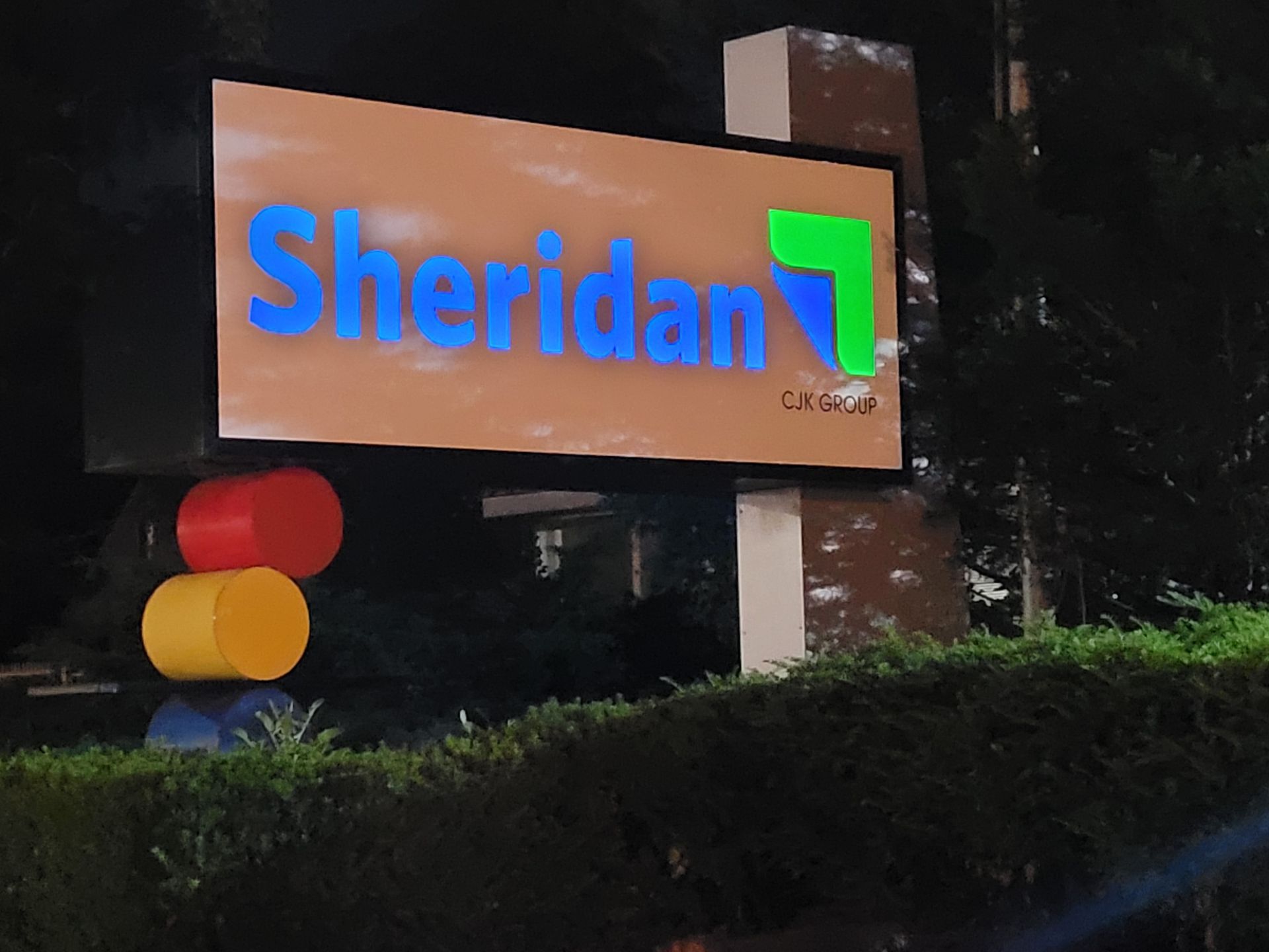 Sheridan campus sign at night with blue and green logo above a red-yellow light post marker