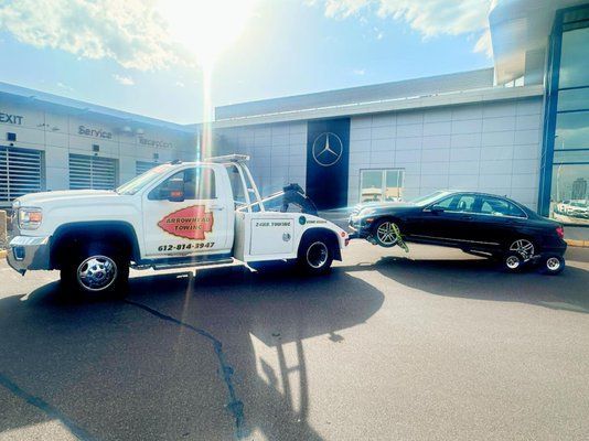 A white tow truck is towing a black car in front of a mercedes dealership.