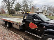 A black tow truck with a flat bed is parked on the side of the road.