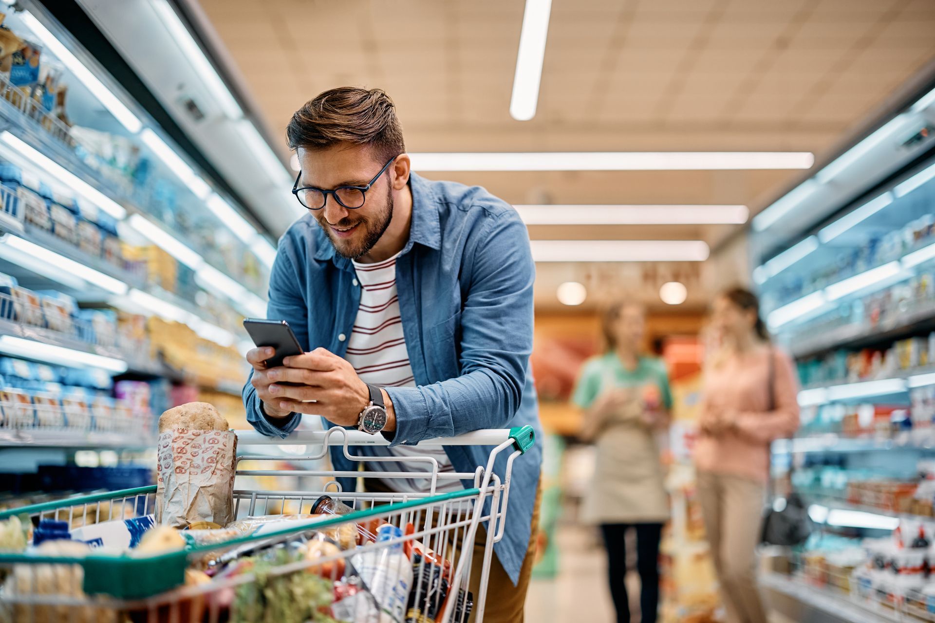 Man in a blue shirt smiling at his phone while grocery shopping. His cart is full of products.