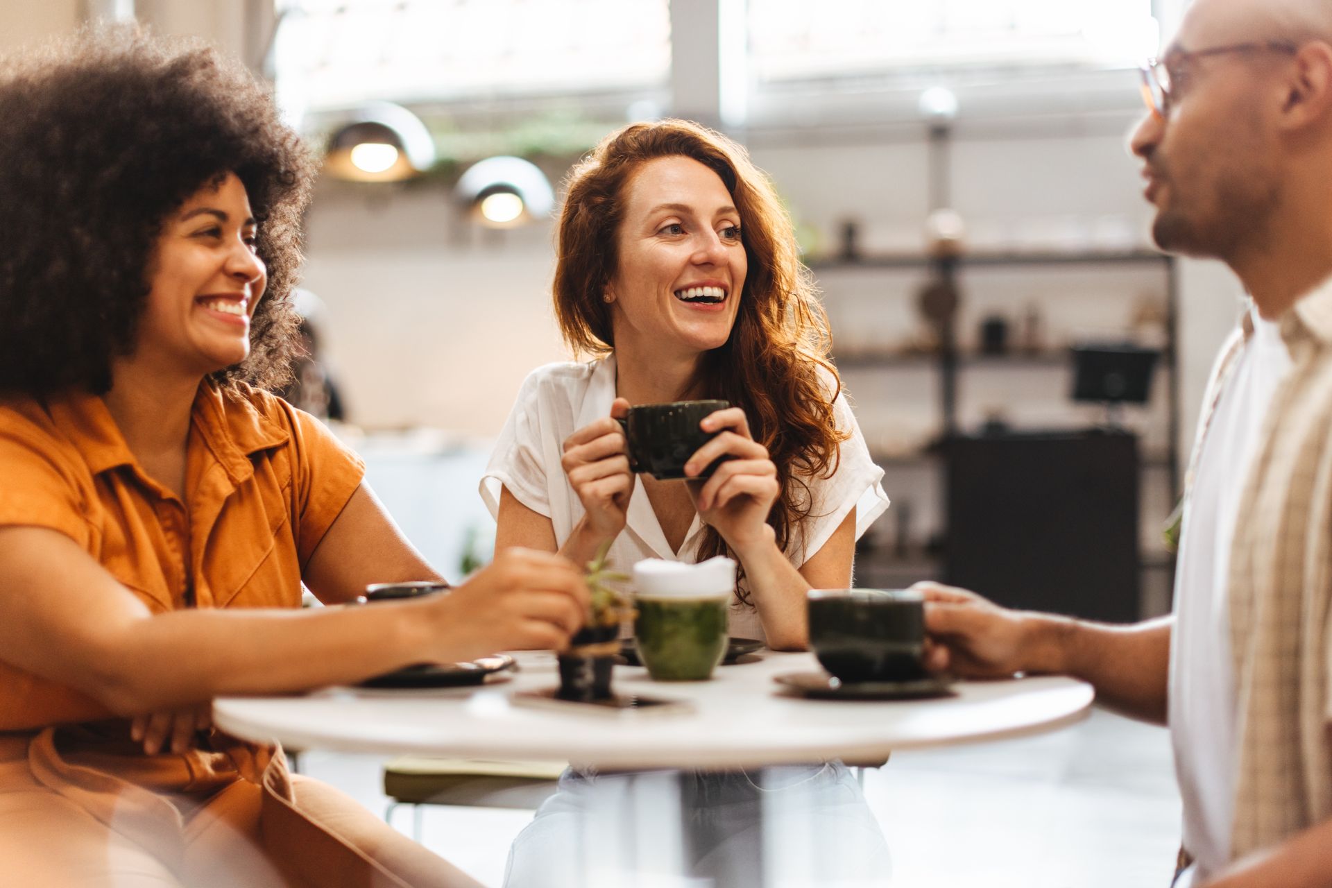 Three friends laughing together, drinking coffee at a cafe.