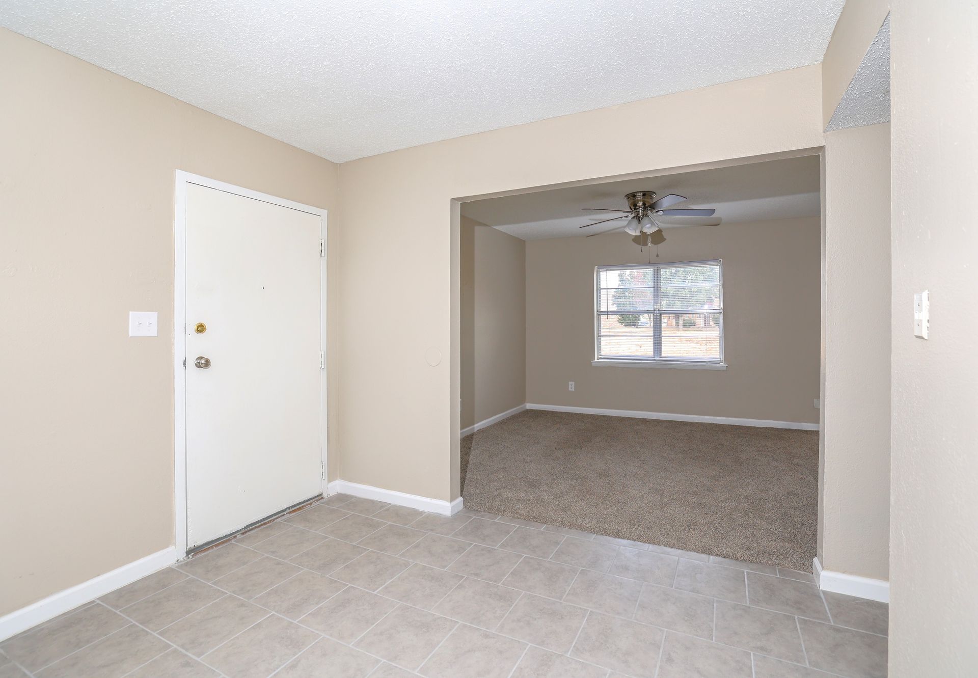 Beige-walled entryway with white door and tile floor, opening to carpeted living room with a window and ceiling fan.