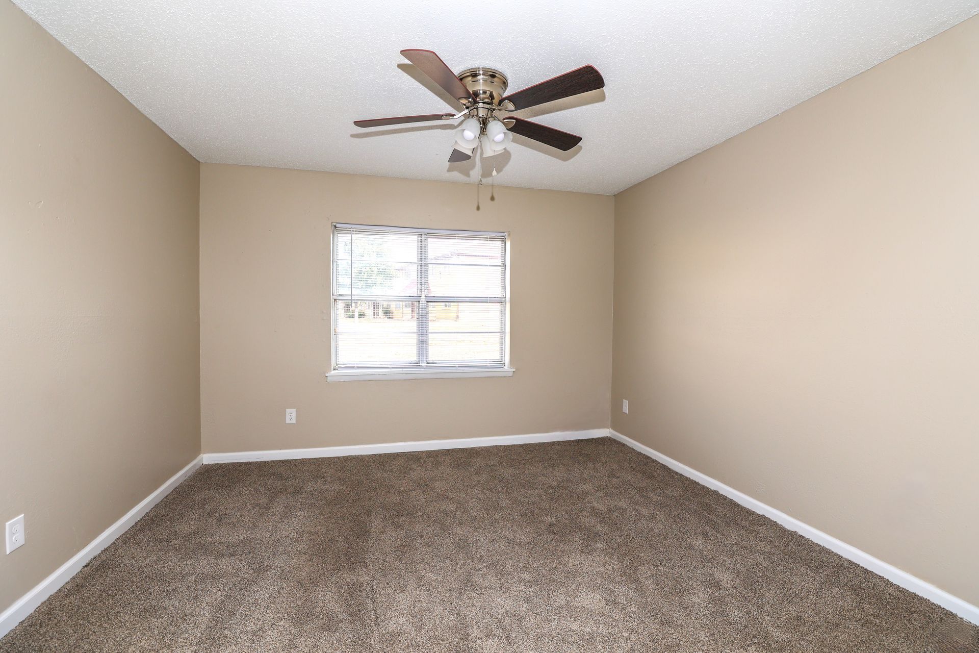 Empty bedroom with beige walls, carpet, window, and ceiling fan.