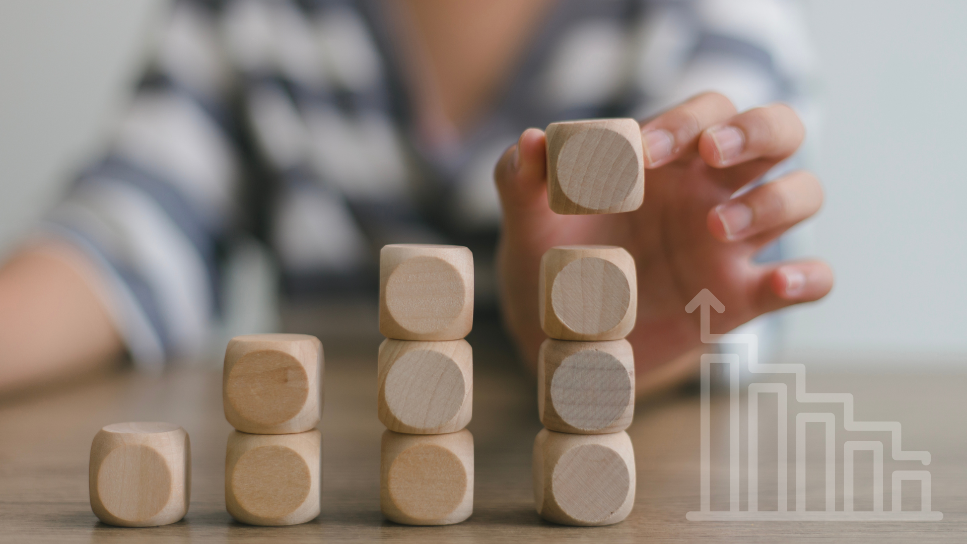 Hand placing a wooden cube atop a stack, next to a rising graph, indicating growth.