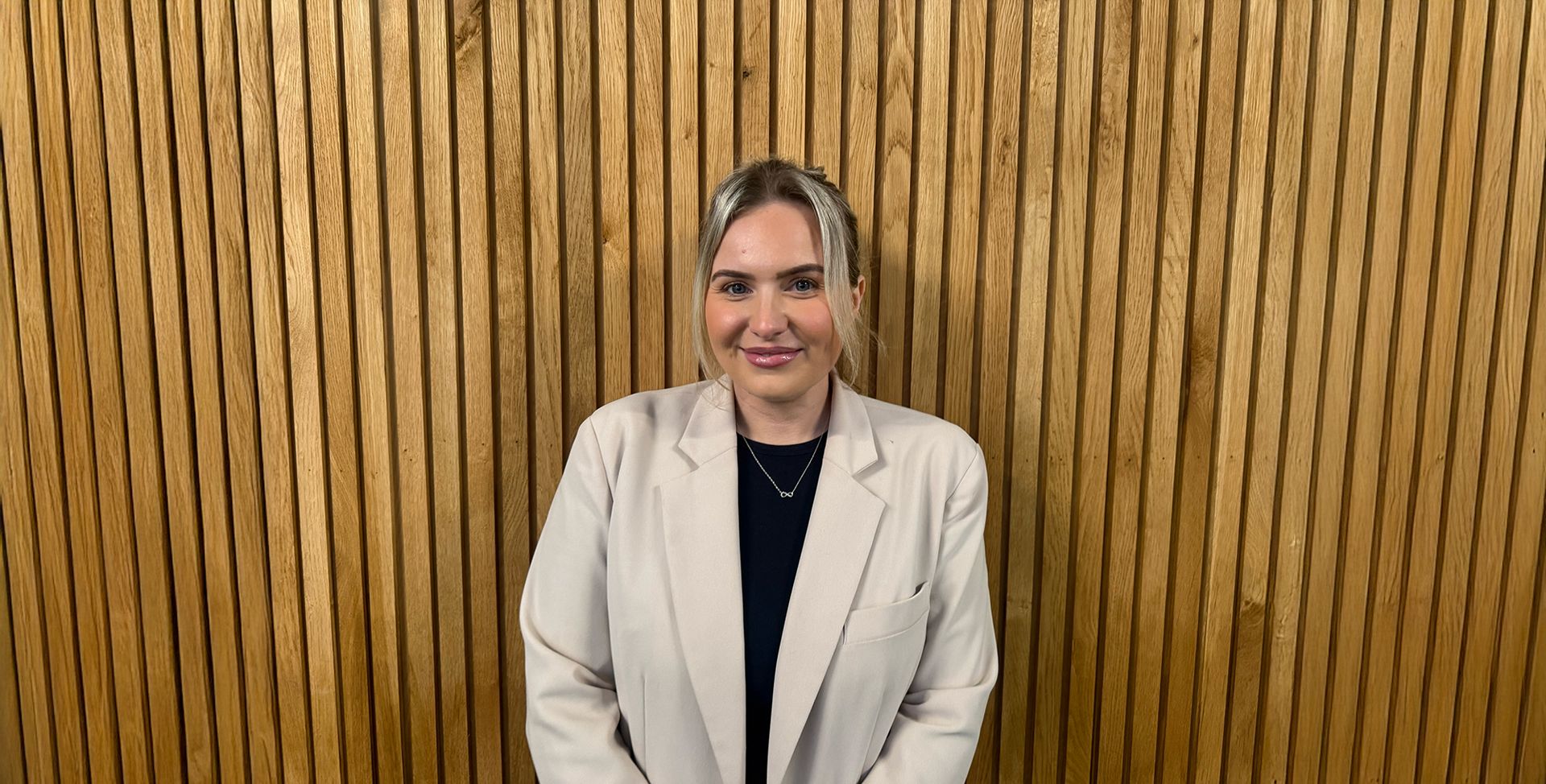 Woman in a cream blazer and dark shirt smiles in front of a wood-paneled wall.