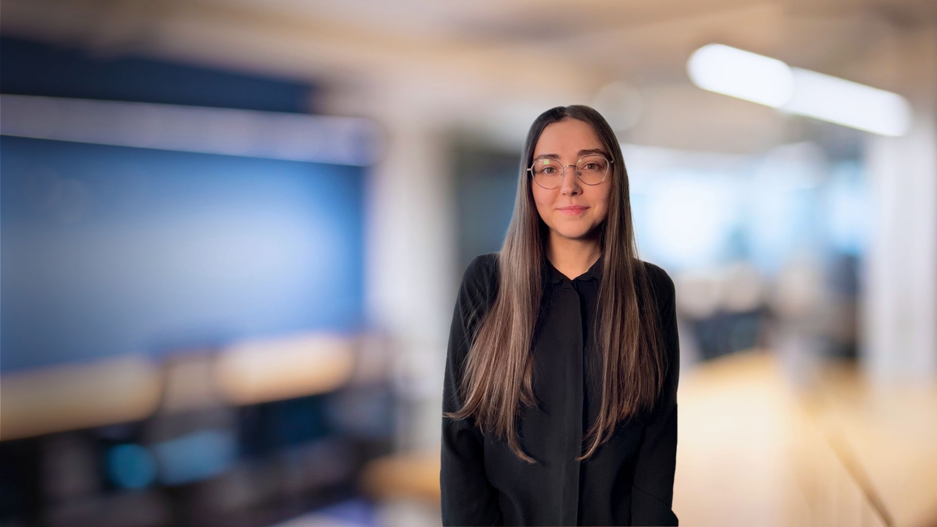 Woman with long brown hair wearing glasses and a black shirt, smiling in an office setting.