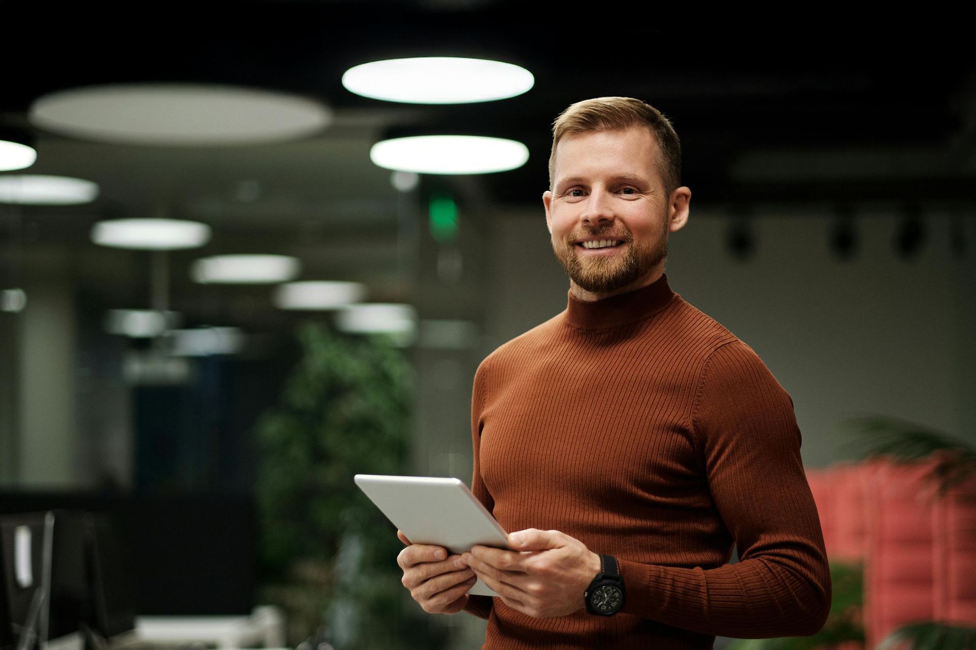 Man in brown turtleneck holding tablet, smiling in office.
