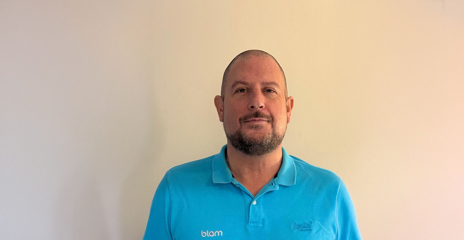 Man in turquoise shirt smiles at camera against a cream-colored wall.