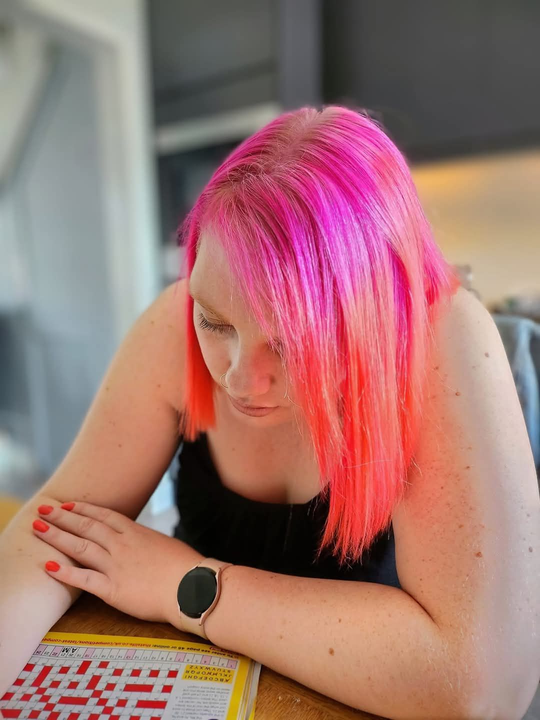 A woman with pink hair is sitting at a table looking at a crossword puzzle.