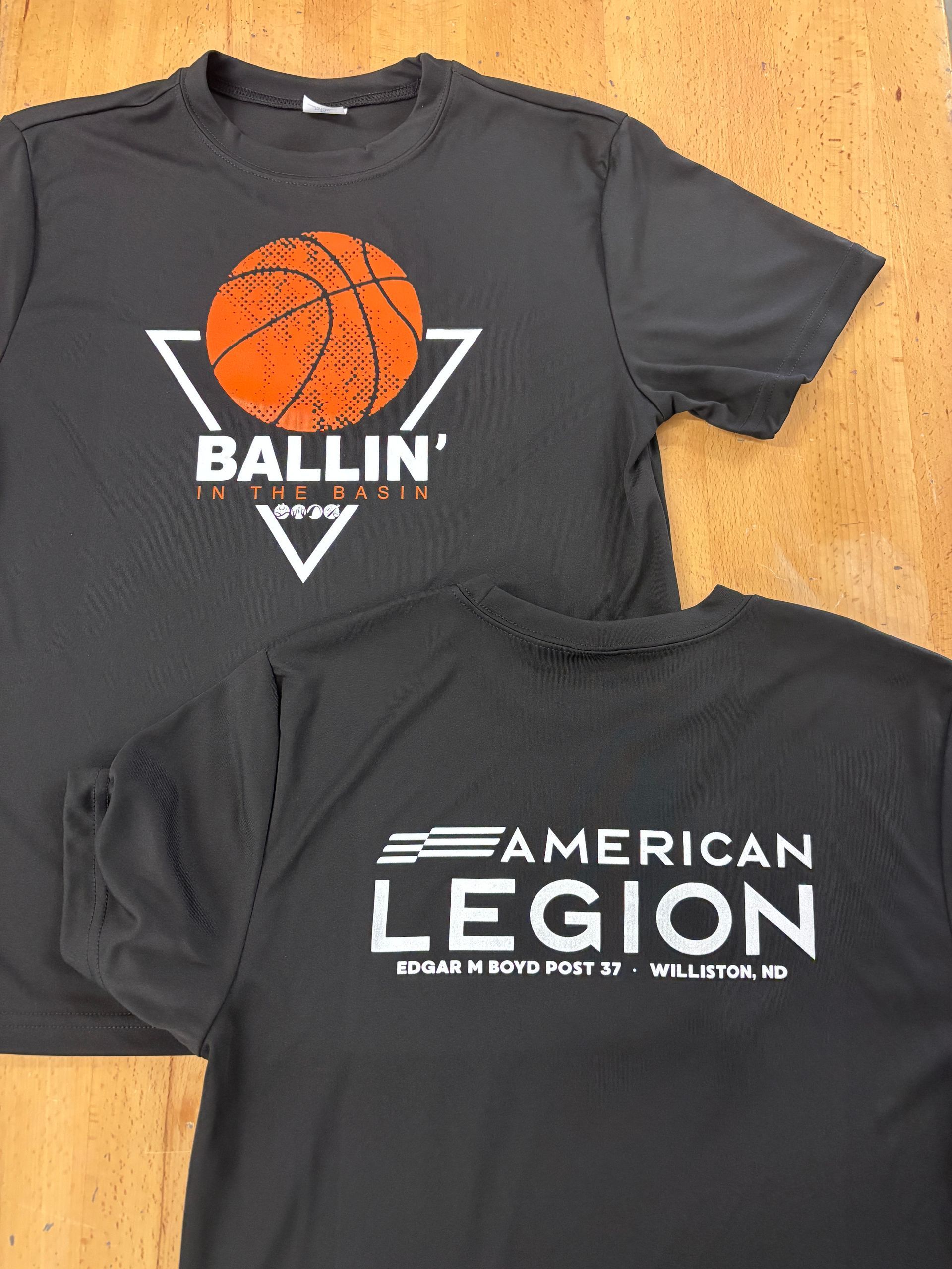 Two black graphic T-shirts laid on a wooden floor, one with a basketball “BALLIN” print and one with “AMERICAN LEGION” text
