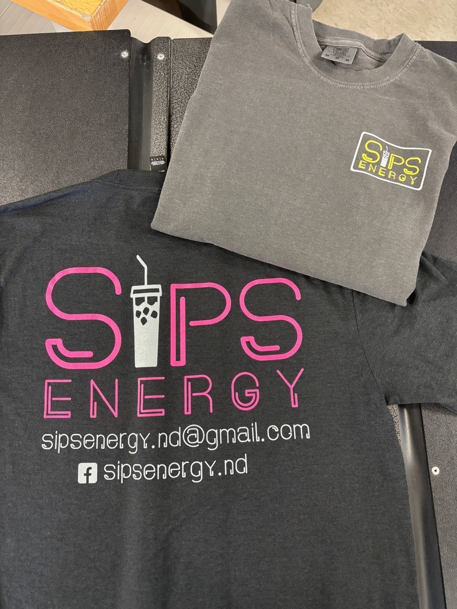 Black SIPS Energy T-shirt with pink logo and contact text, plus a gray folded shirt on top