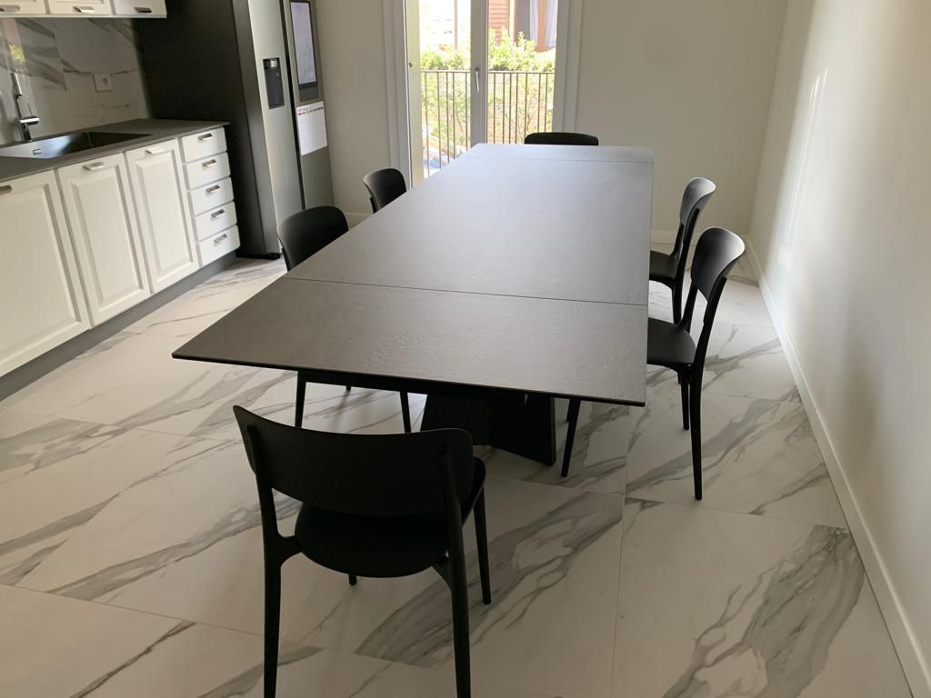 Sala da pranzo con un lungo tavolo nero e sei sedie nere, pavimento in piastrelle chiare.