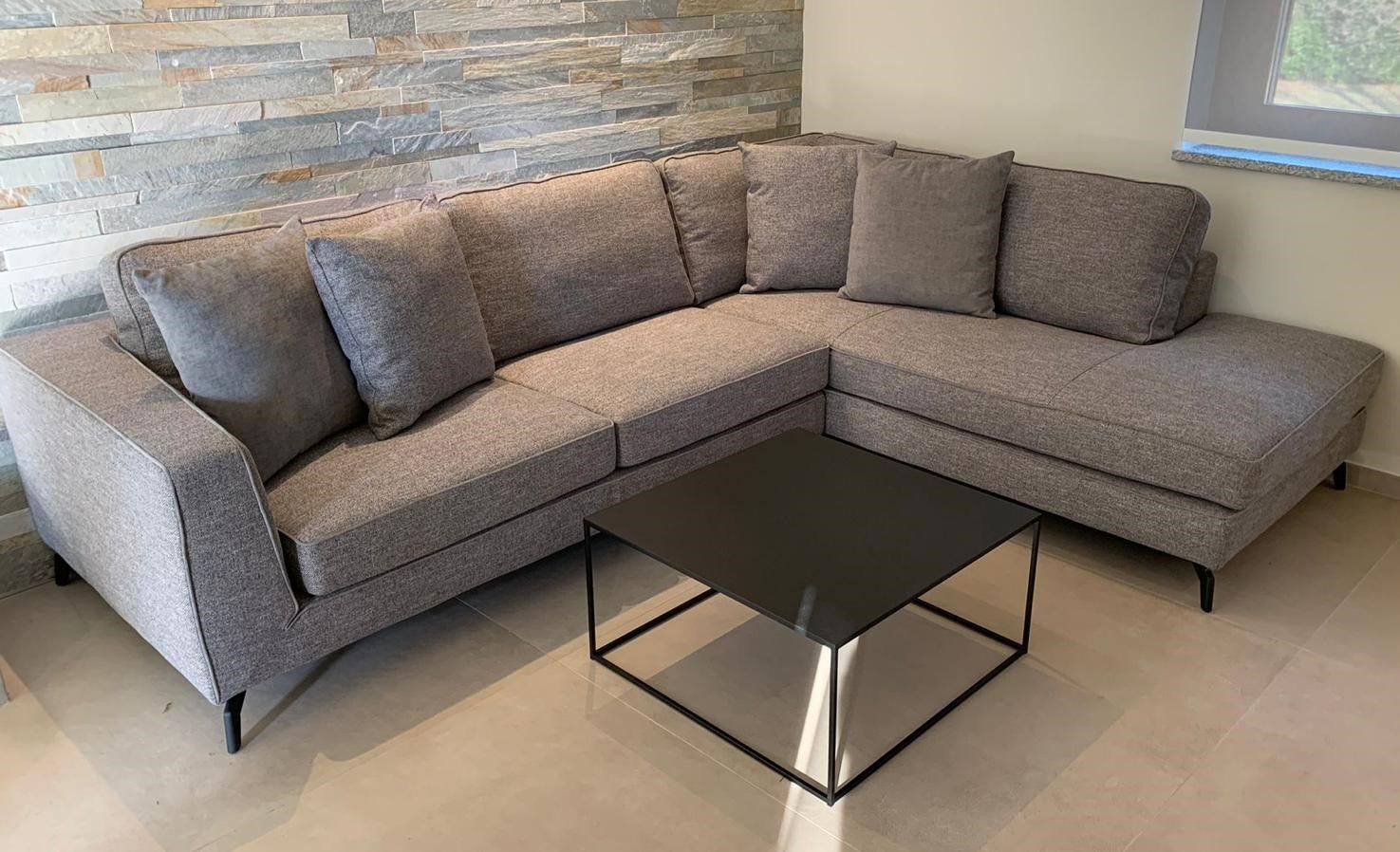 Divano componibile grigio con cuscini, tavolino quadrato nero, parete in pietra sullo sfondo.