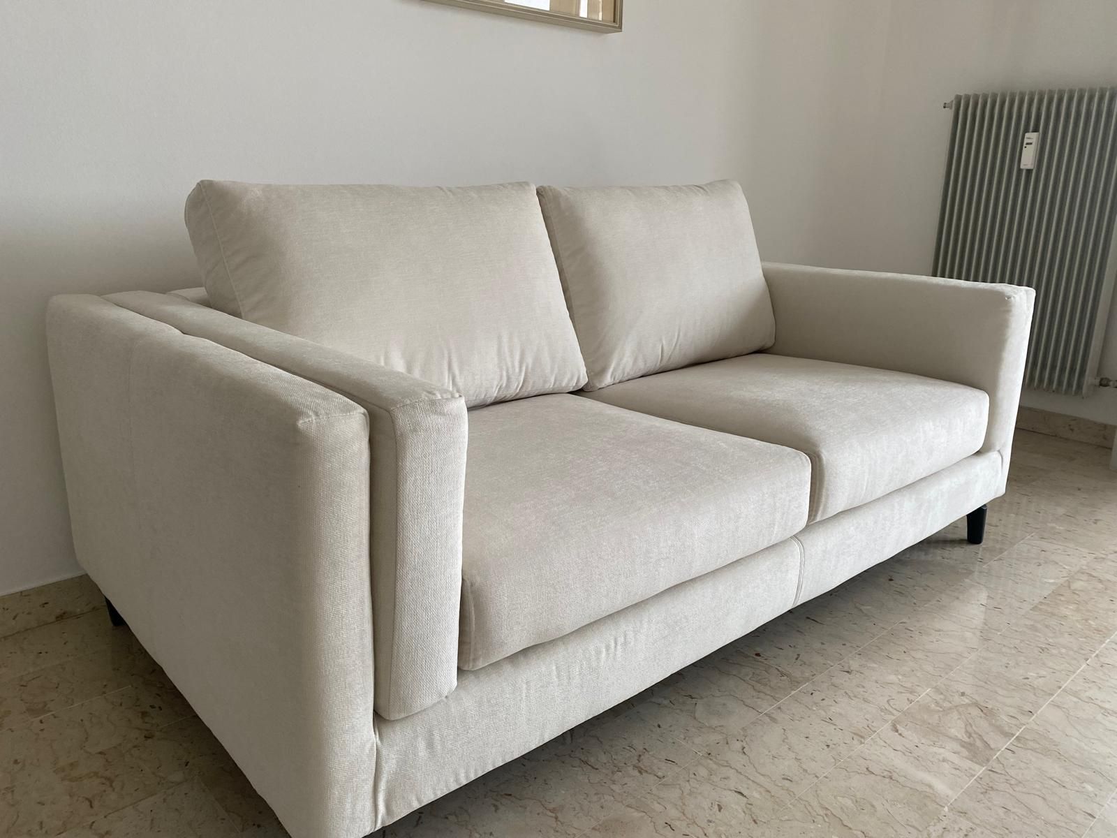 Divano beige a due posti con braccioli quadrati e cuscini, posizionato di fronte a una parete con un termosifone.