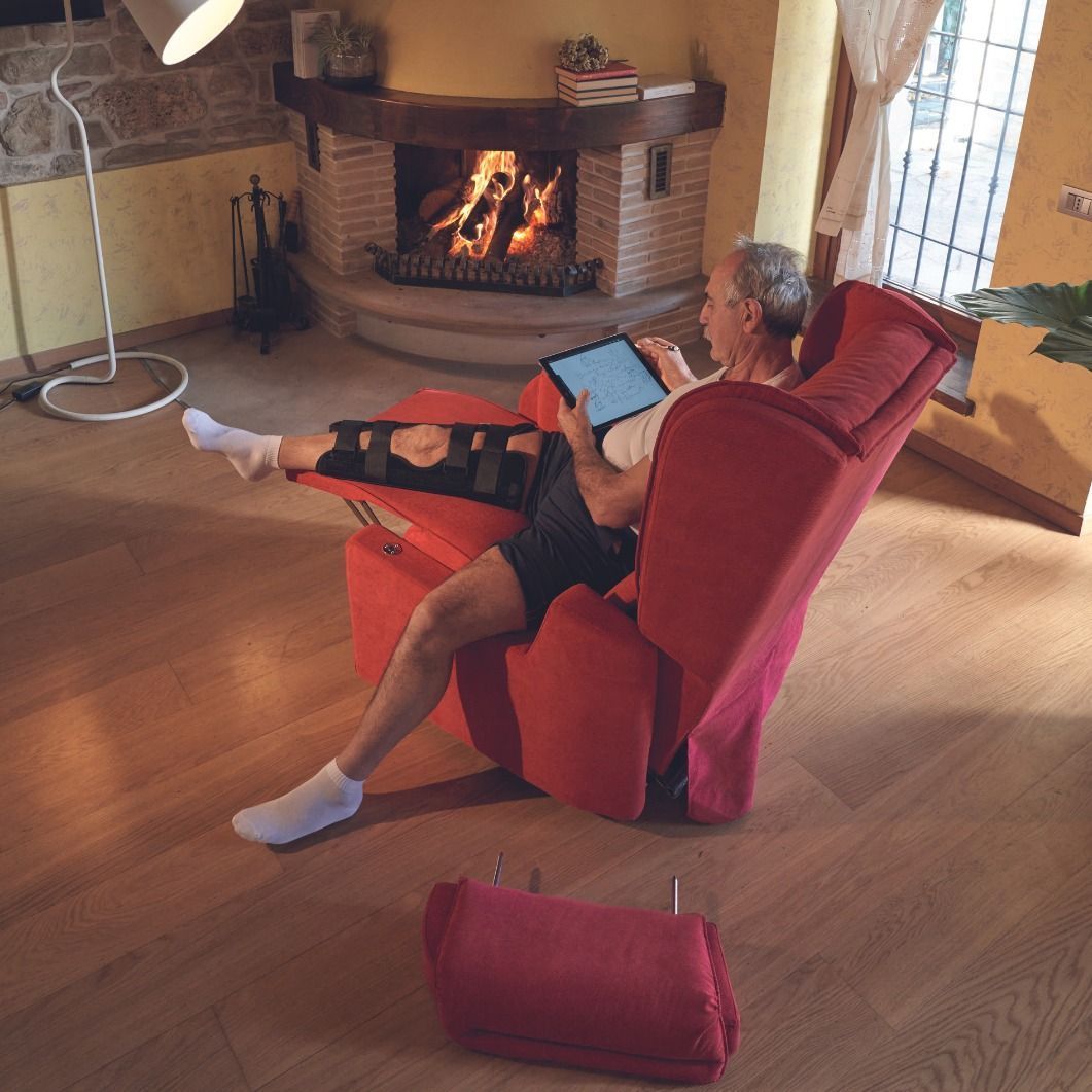 Un uomo con un tutore per la gamba si rilassa su una poltrona reclinabile rossa, usando un tablet, vicino al camino.