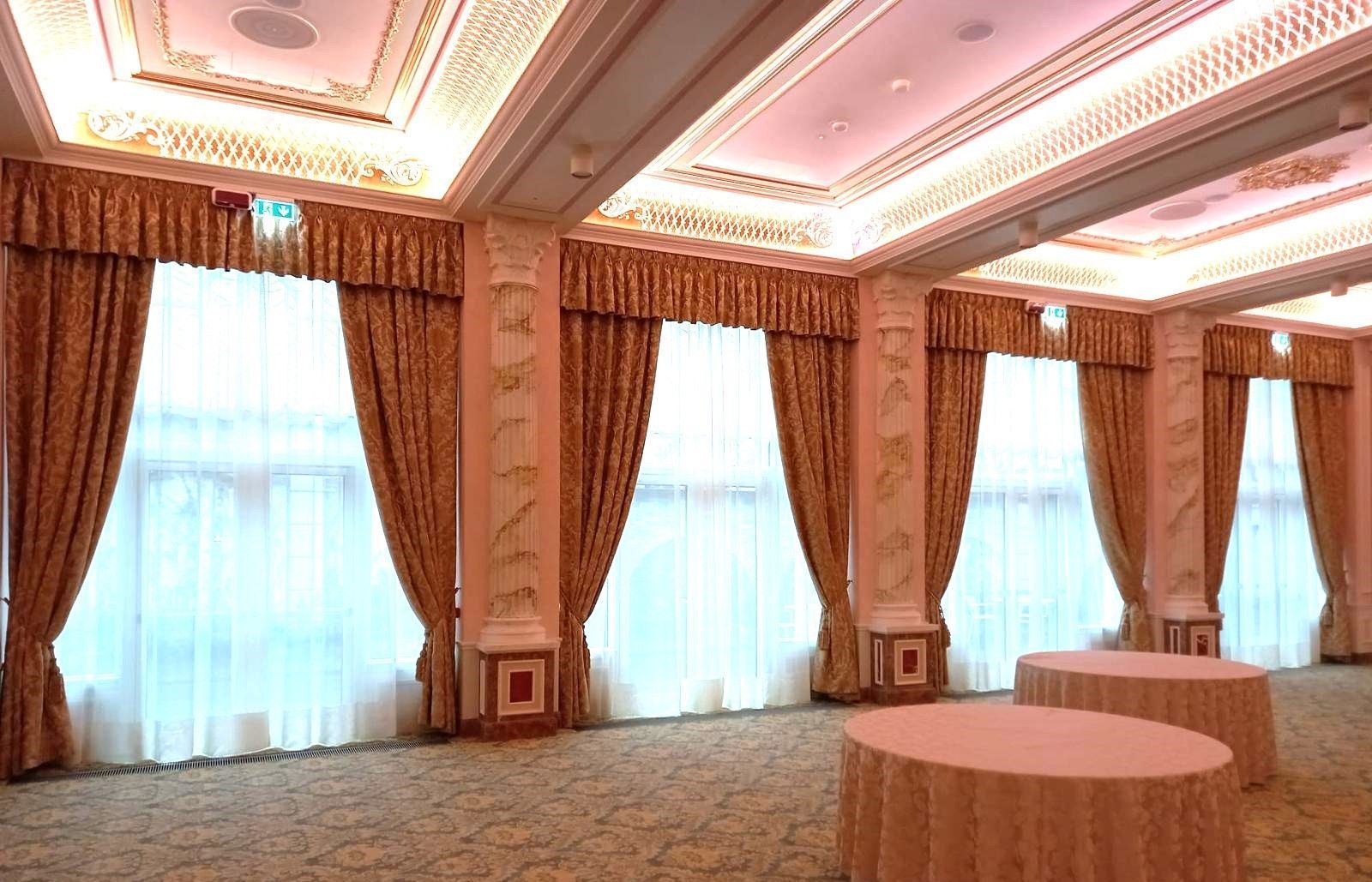 Elegante sala da ballo con soffitto decorato, grandi finestre con tende dorate e tavoli rotondi.