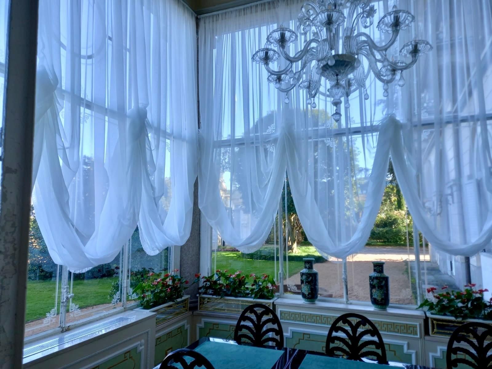 Stanza con tende bianche trasparenti, ampie finestre, lampadario e tavolo apparecchiato.