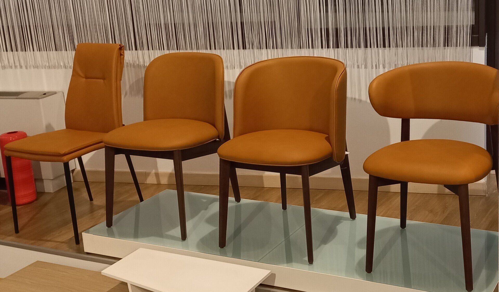Quattro sedie in pelle arancione con gambe scure su una piattaforma bianca, contro una parete decorata.