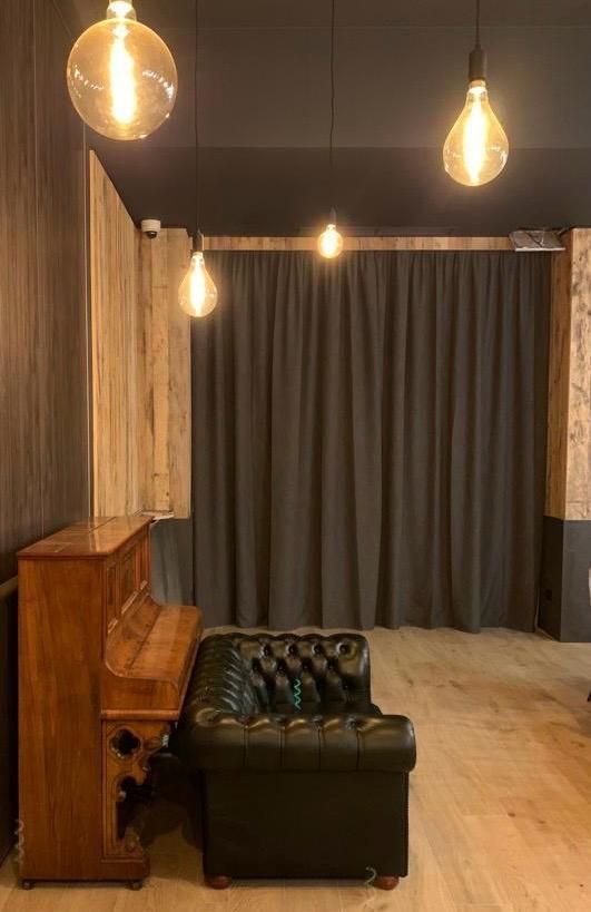 Un pianoforte in legno, un divano in pelle verde e luci sospese in una stanza con pannelli in legno e tende scure.