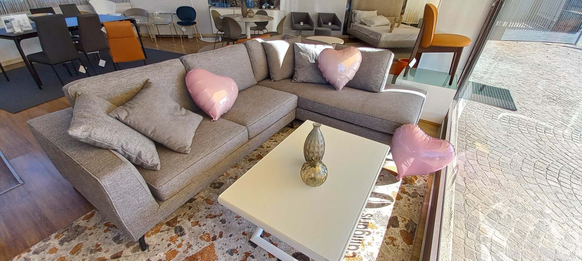 Divano componibile grigio con cuscini rosa a forma di cuore, un tavolino bianco e un vaso decorativo in un negozio di mobili.