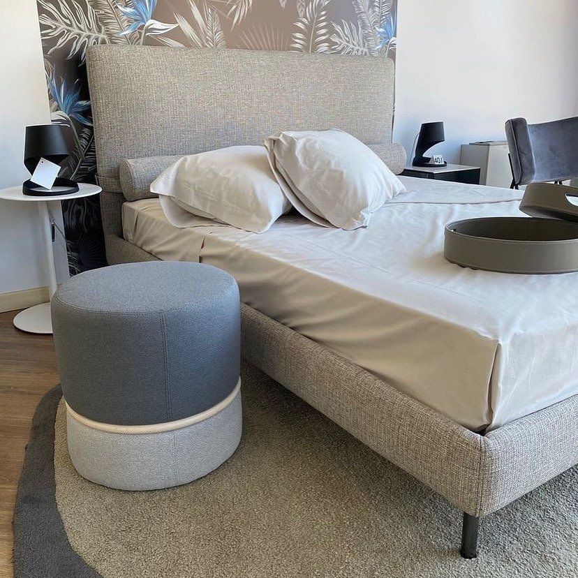 Scena di camera da letto con letto, pouf, comodino e tappeto. Dominano i toni del grigio e del beige.