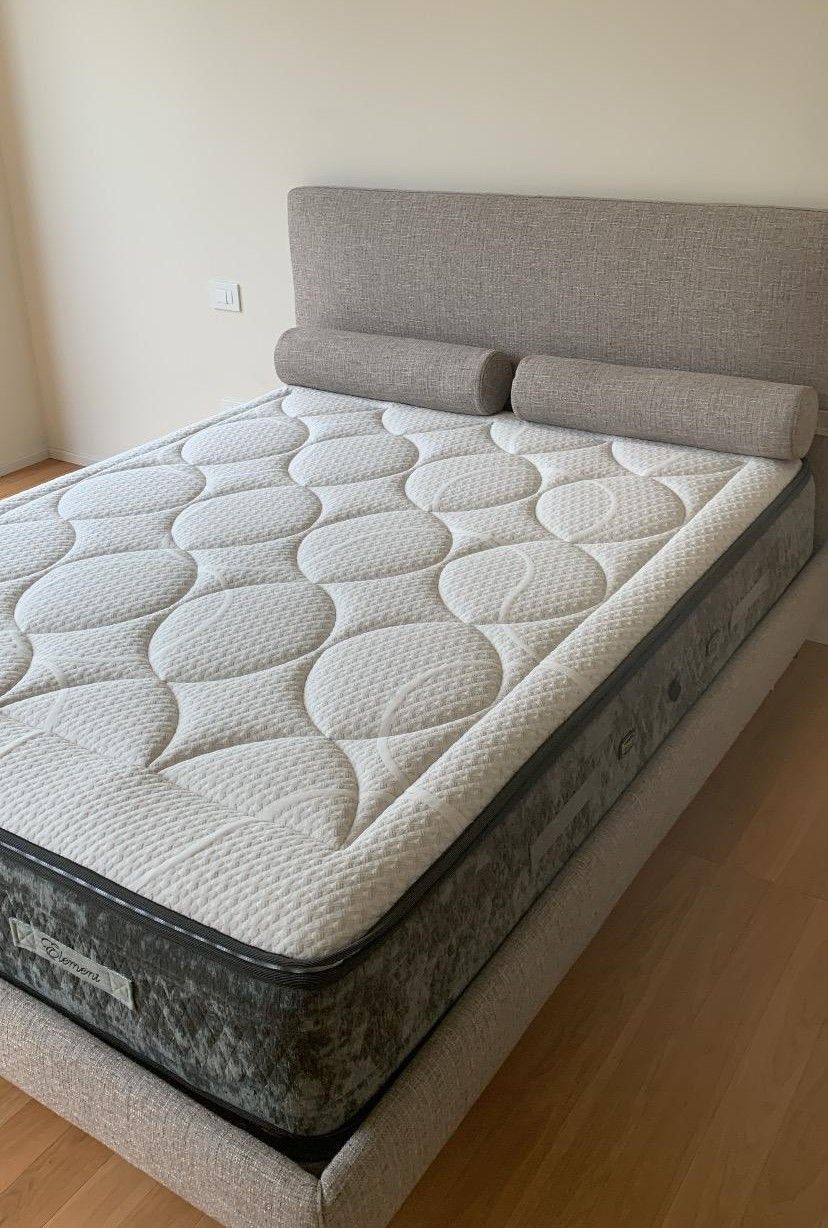 Letto imbottito con testiera e materasso grigi. Sul letto sono appoggiati due cuscini cilindrici.