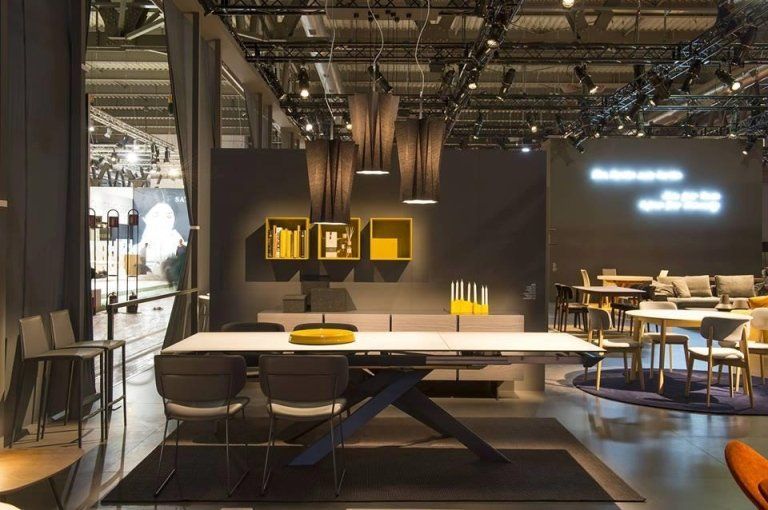 Esposizione di mobili moderni con un grande tavolo, sedie, scaffali e illuminazione. Toni del grigio e del giallo.