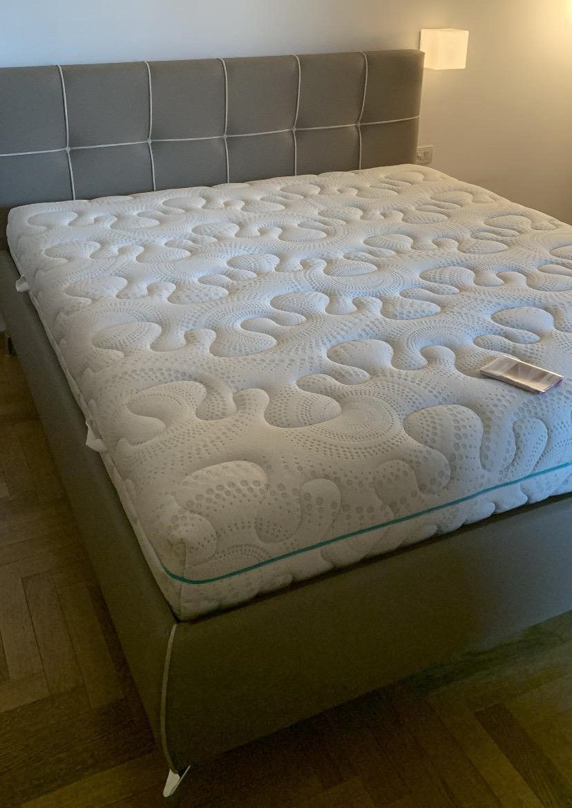 Letto imbottito grigio con materasso e testiera con fantasia bianca, su pavimento in legno.