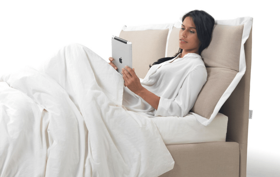 Donna a letto che legge un tablet, appoggiata ai cuscini. Lenzuola e testiera beige.