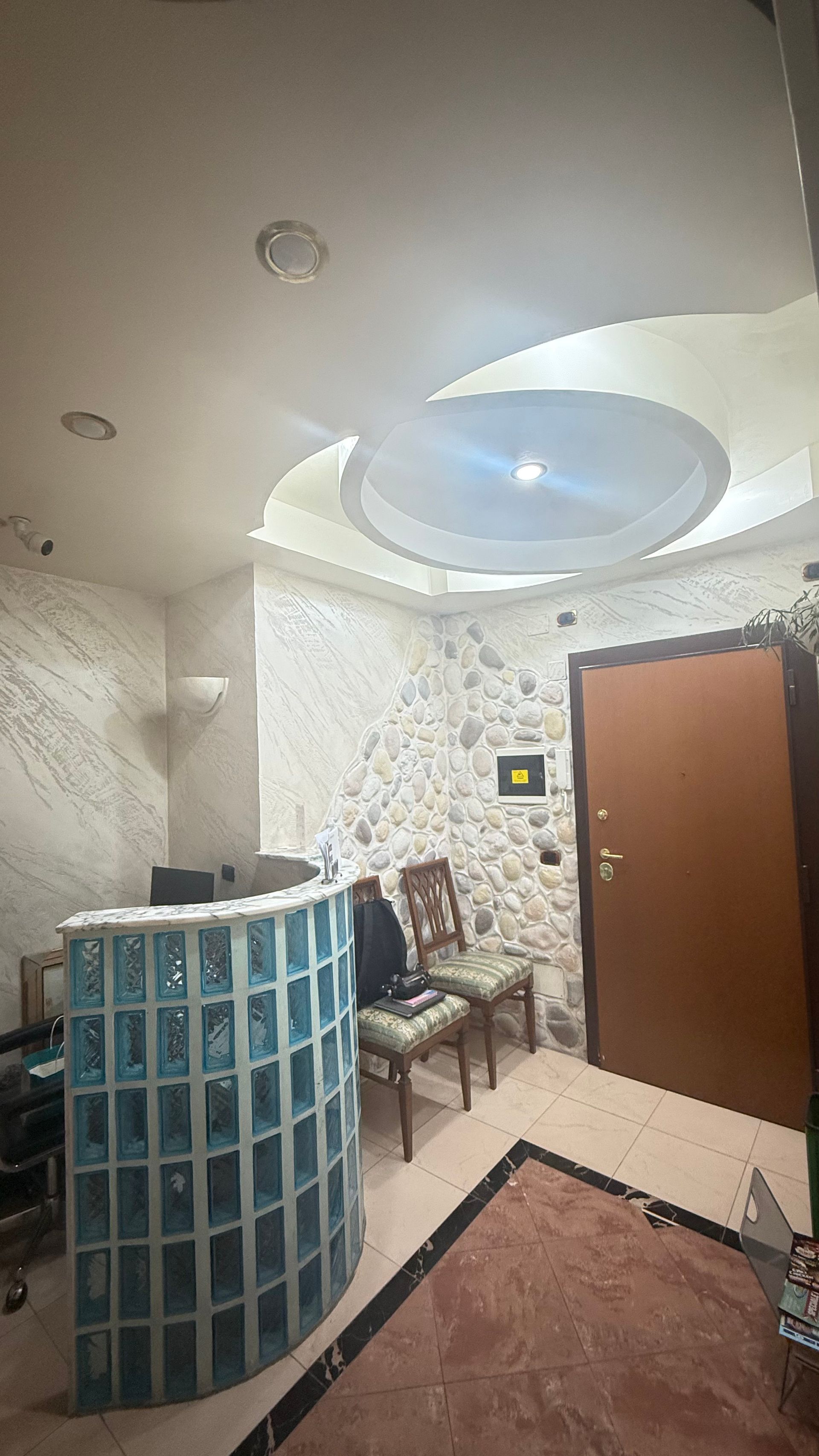 Area reception: scrivania in vetrocemento blu, pareti bianche testurizzate, porta marrone, due sedie, soffitto decorativo.