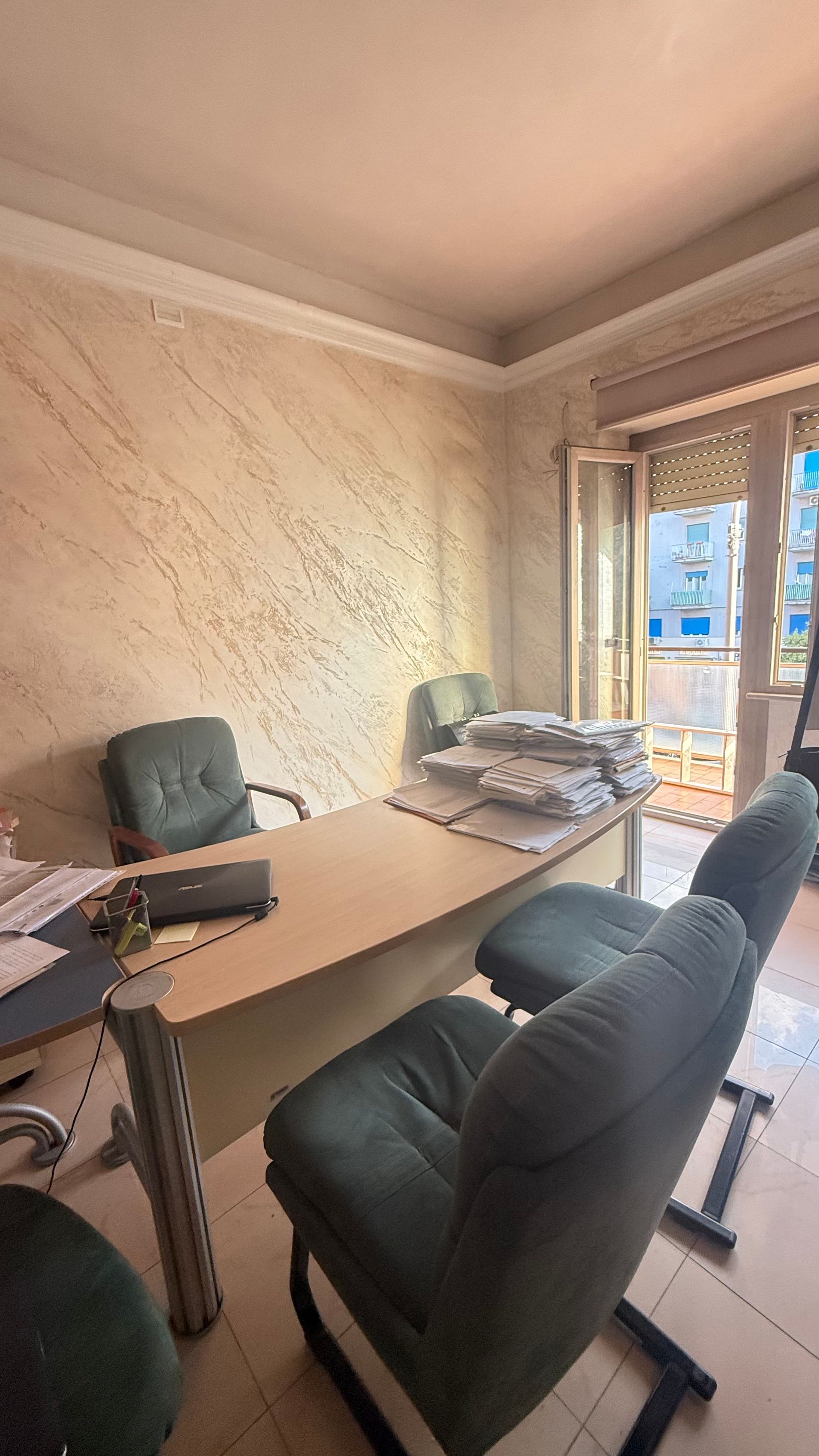 Interno di un ufficio con scrivania, sedie e balcone. Pareti color crema e una luminosa vista esterna.