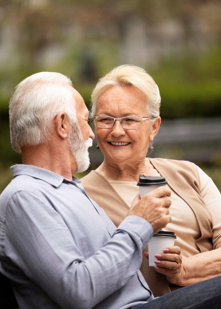 An elderly couple is sitting on a park bench drinking coffee.