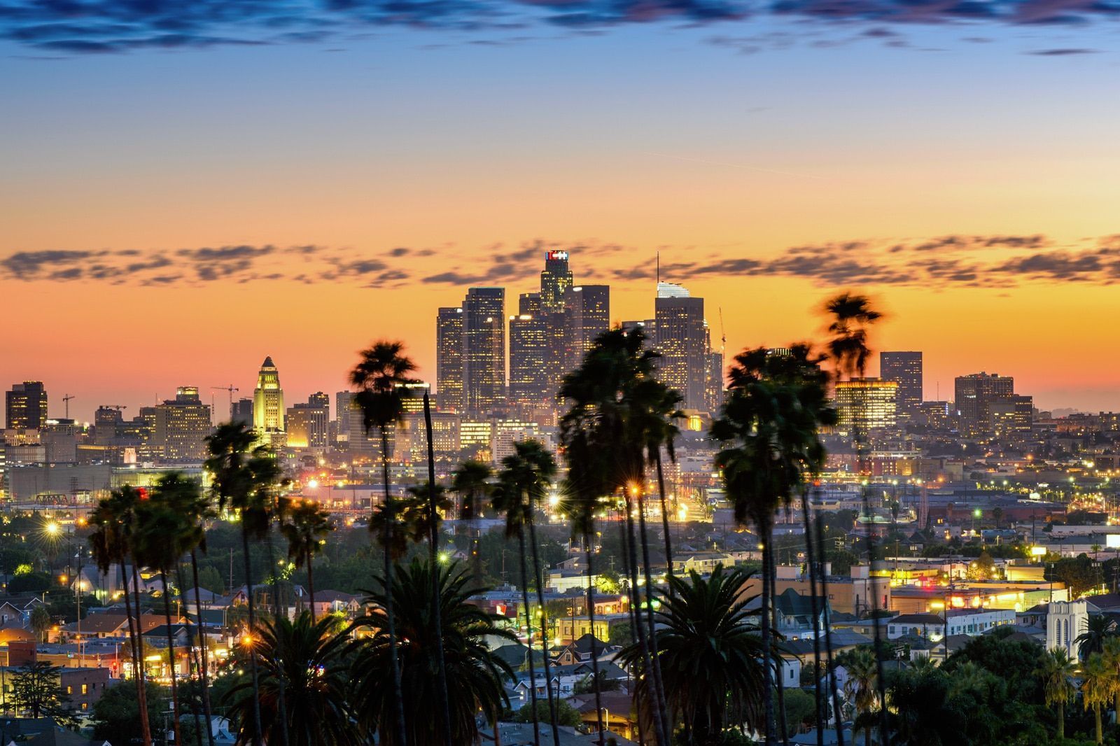 Downtown Los Angeles skyline at dusk with palm trees in the foreground and an orange and blue sky.