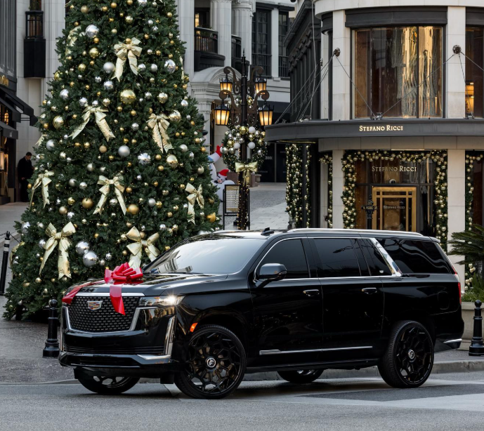 Black Cadillac SUV with a red bow parked in front of a Christmas tree in a city setting.