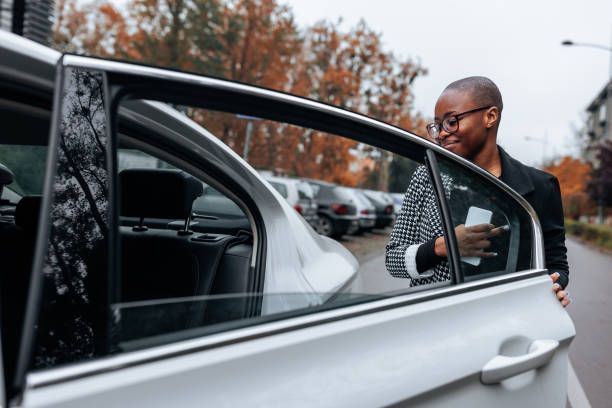 Woman in blazer opens a car door, holding a phone. Outdoors, cars parked in background.
