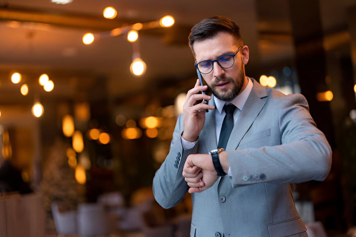 Man in a suit looks at his watch while talking on a phone in a dimly lit restaurant.