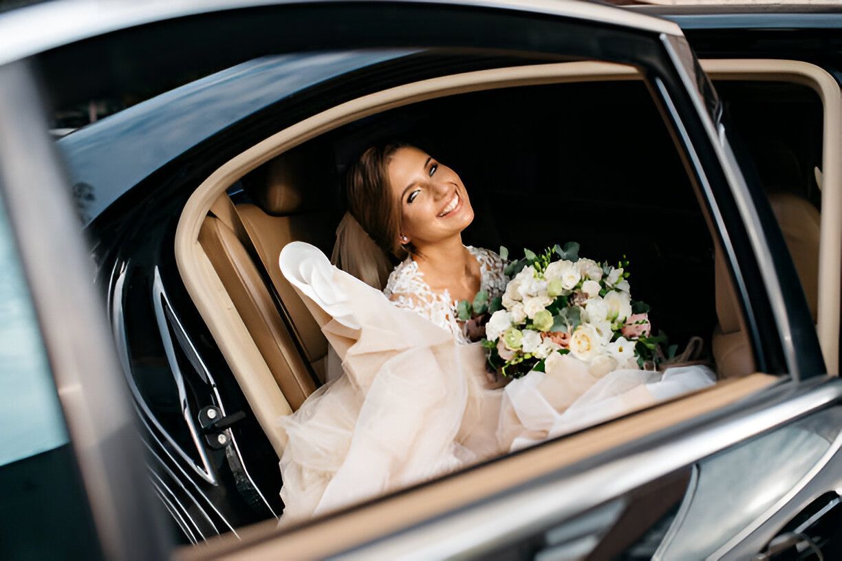 Bride in wedding dress smiles, holding bouquet, seated in a car.