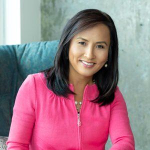 A woman in a pink jacket is sitting in a chair and smiling.