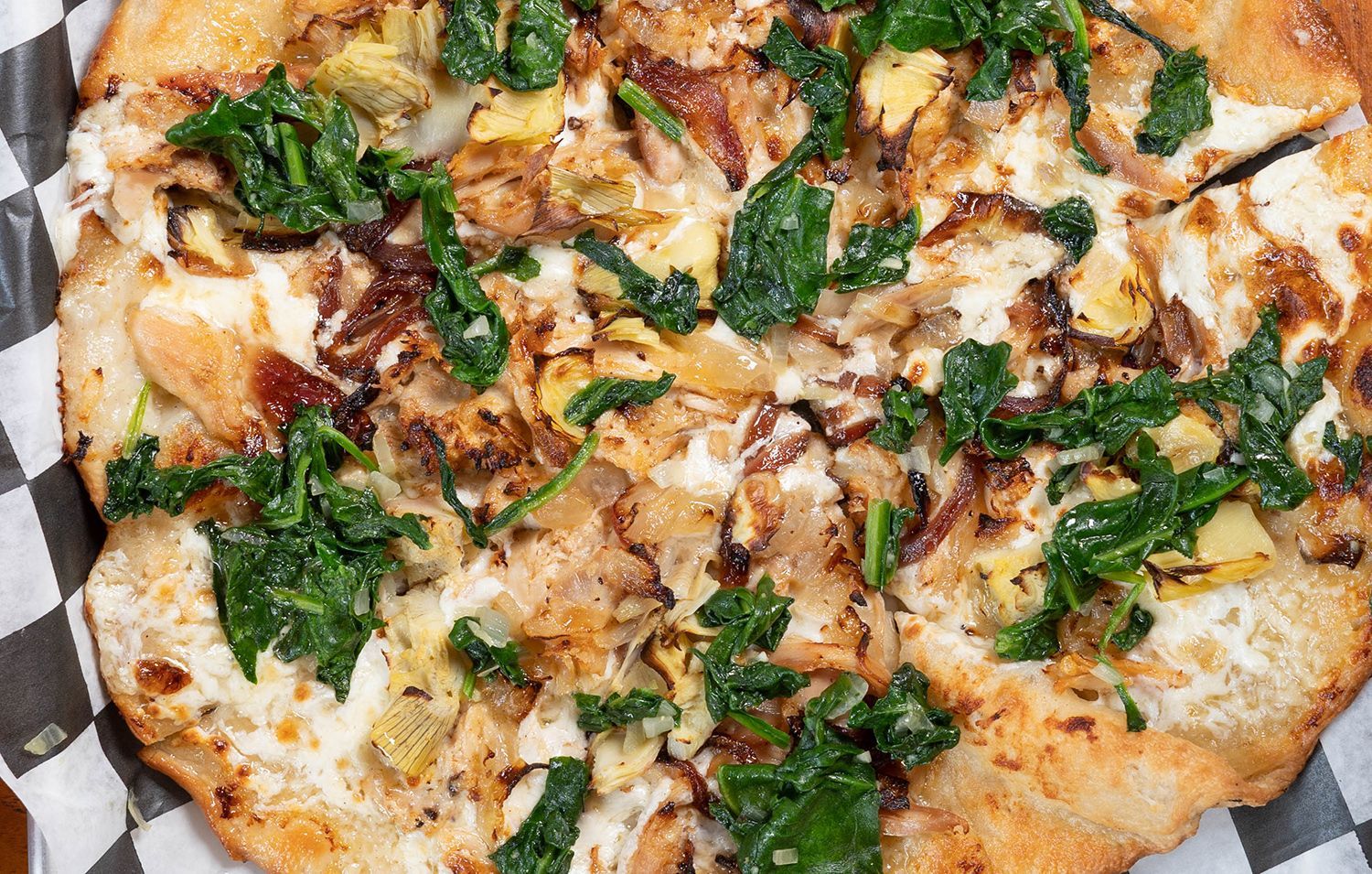 A pizza with spinach , artichokes , onions and cheese on a checkered napkin.