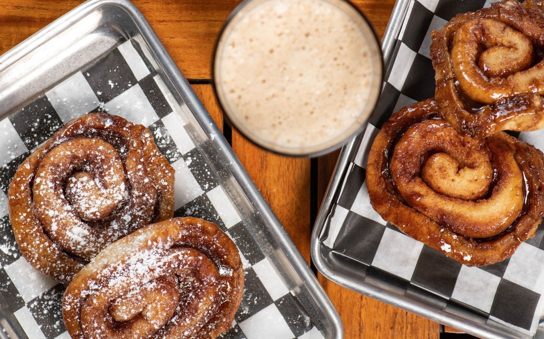 Two trays of cinnamon rolls and a glass of milk on a wooden table.