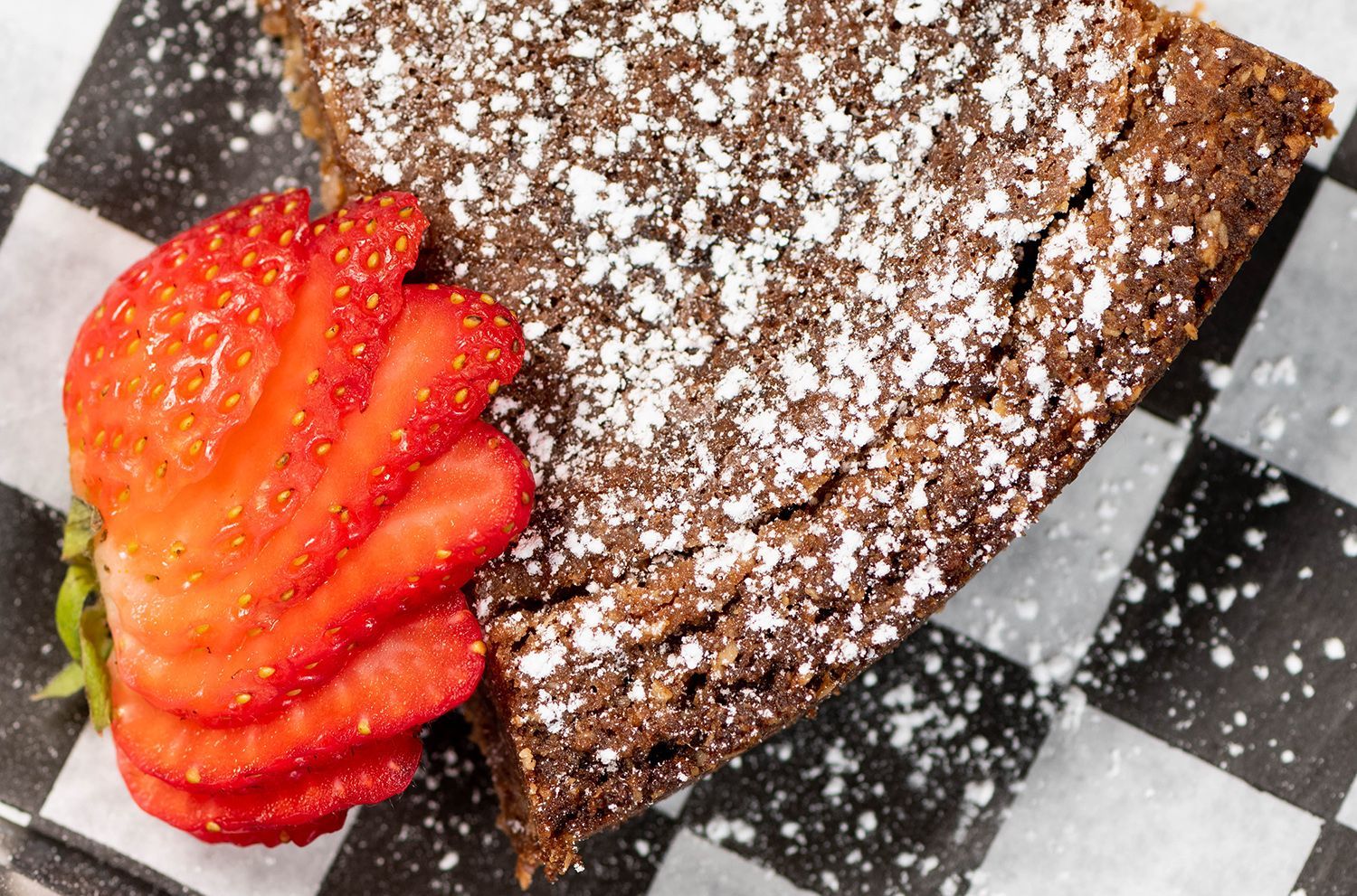 A piece of chocolate cake with powdered sugar and strawberries on a checkered napkin.