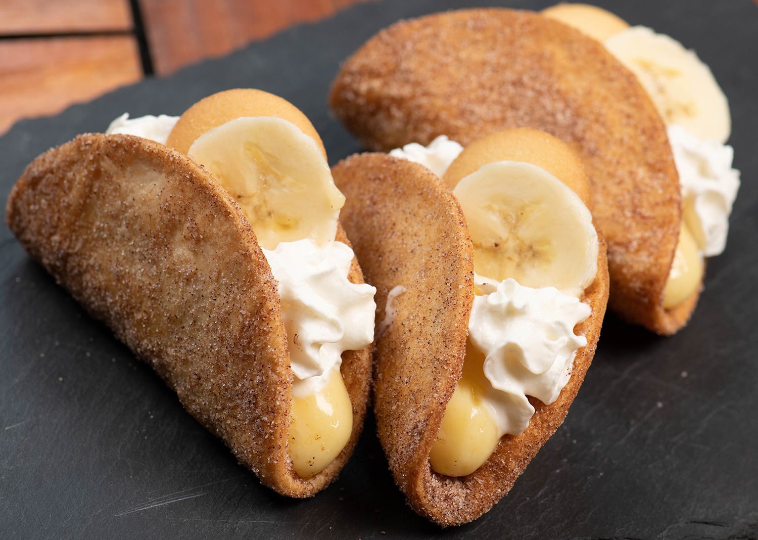 Three banana tacos with whipped cream and banana slices on a black plate.