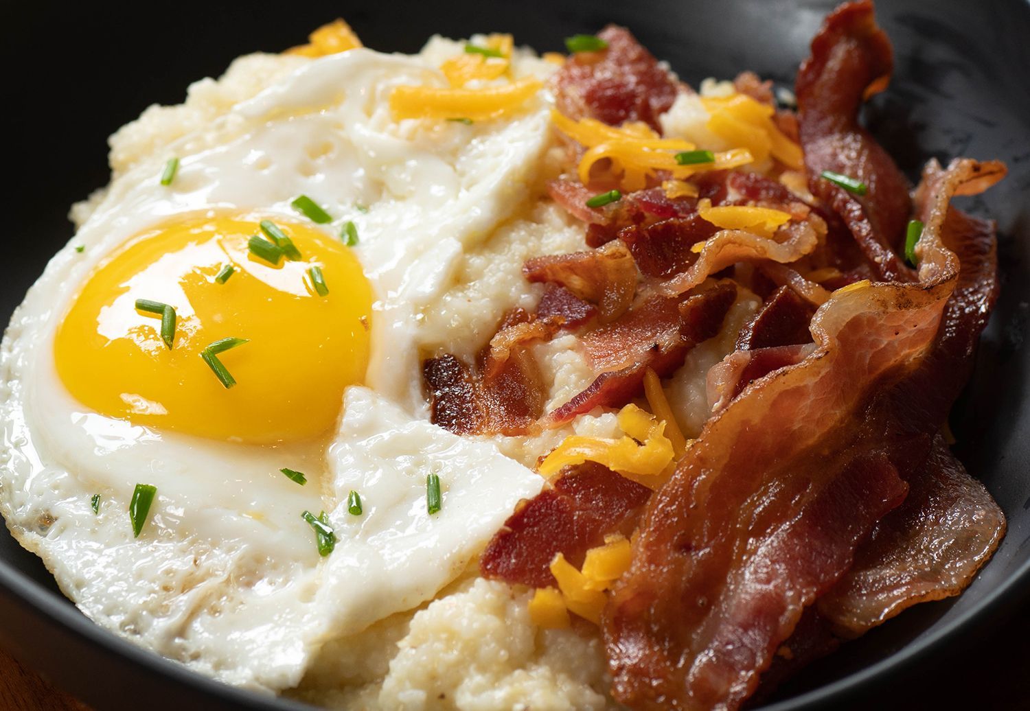 A close up of a bowl of food with eggs , bacon , cheese and grits.
